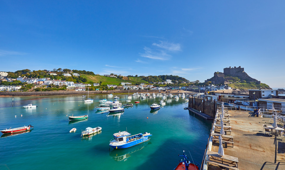 Gorey Harbour with fishing and pleasure boats, the pier bullworks and Gorey Castle in the background with blue sky. Jersey, Channel Islands, UK